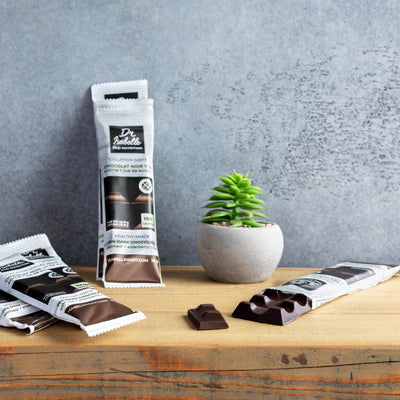 Photographie de carrés de chocolats noir à l'avoine empilés les uns sur les autres sur le premier plan, et en second plan plusieurs tablettes de chocolat noir 70% visibles avec le logo de la marque Isabelle Huot Docteure en nutrition. Photograph of stacked chocolate squares above one another in the foreground. In the background there are two chocolate tablets with the logo of the company Isabelle Huot nutritionist doctor