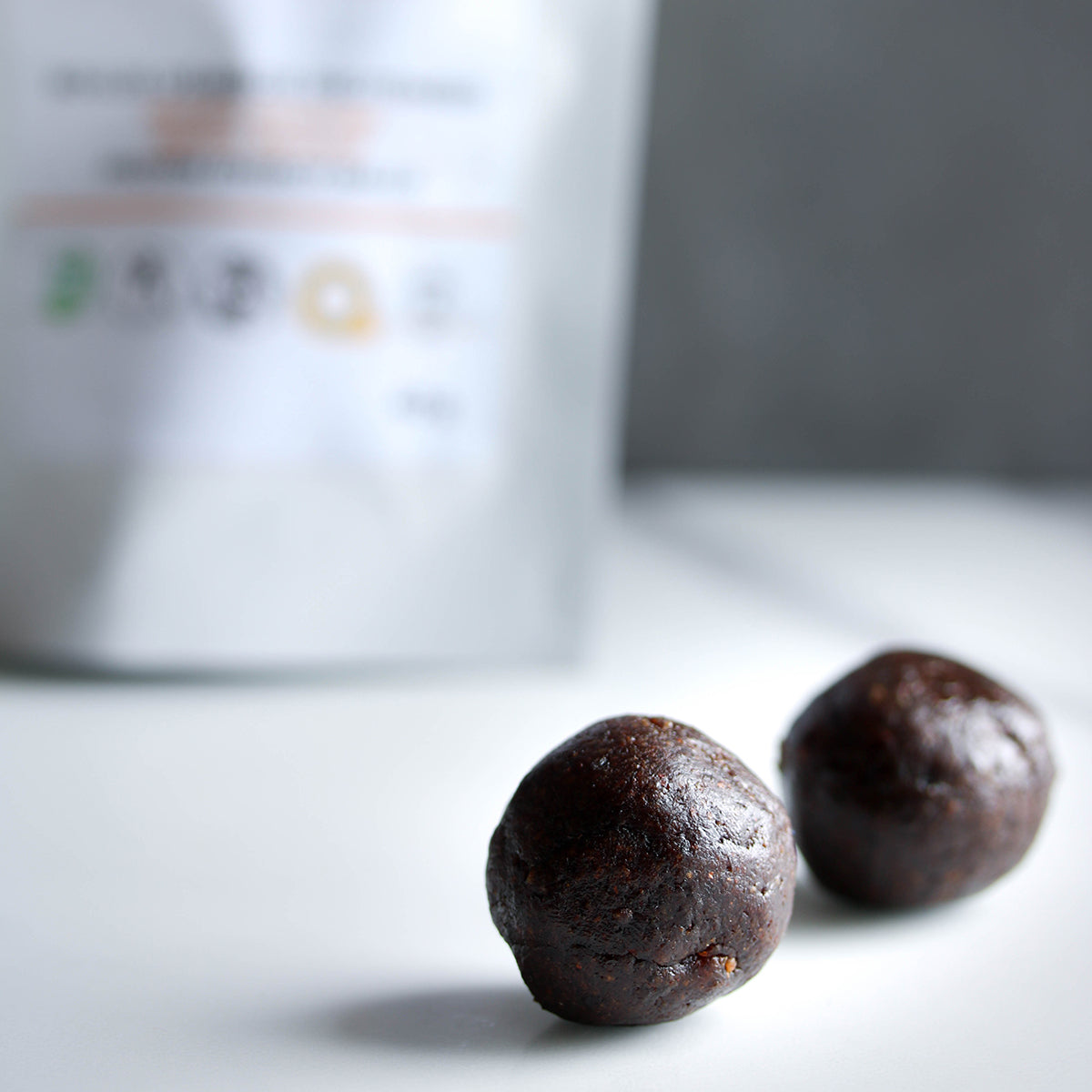 Photographie de deux boules énergie à l'amande et abricot, en premier plan, et leur sachet en second plan. Photograph of two apricot and almond energy balls in the foreground, and their bag in the background. *conçu pour respecter les besoins des personnes atteintes de diabète, d'hypercholestérolémie ou d'hypertension artérielle