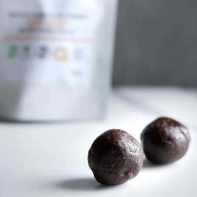 Photographie de deux boules énergie à l'amande et abricot, en premier plan, et leur sachet en second plan. Photograph of two apricot and almond energy balls in the foreground, and their bag in the background. *conçu pour respecter les besoins des personnes atteintes de diabète, d'hypercholestérolémie ou d'hypertension artérielle