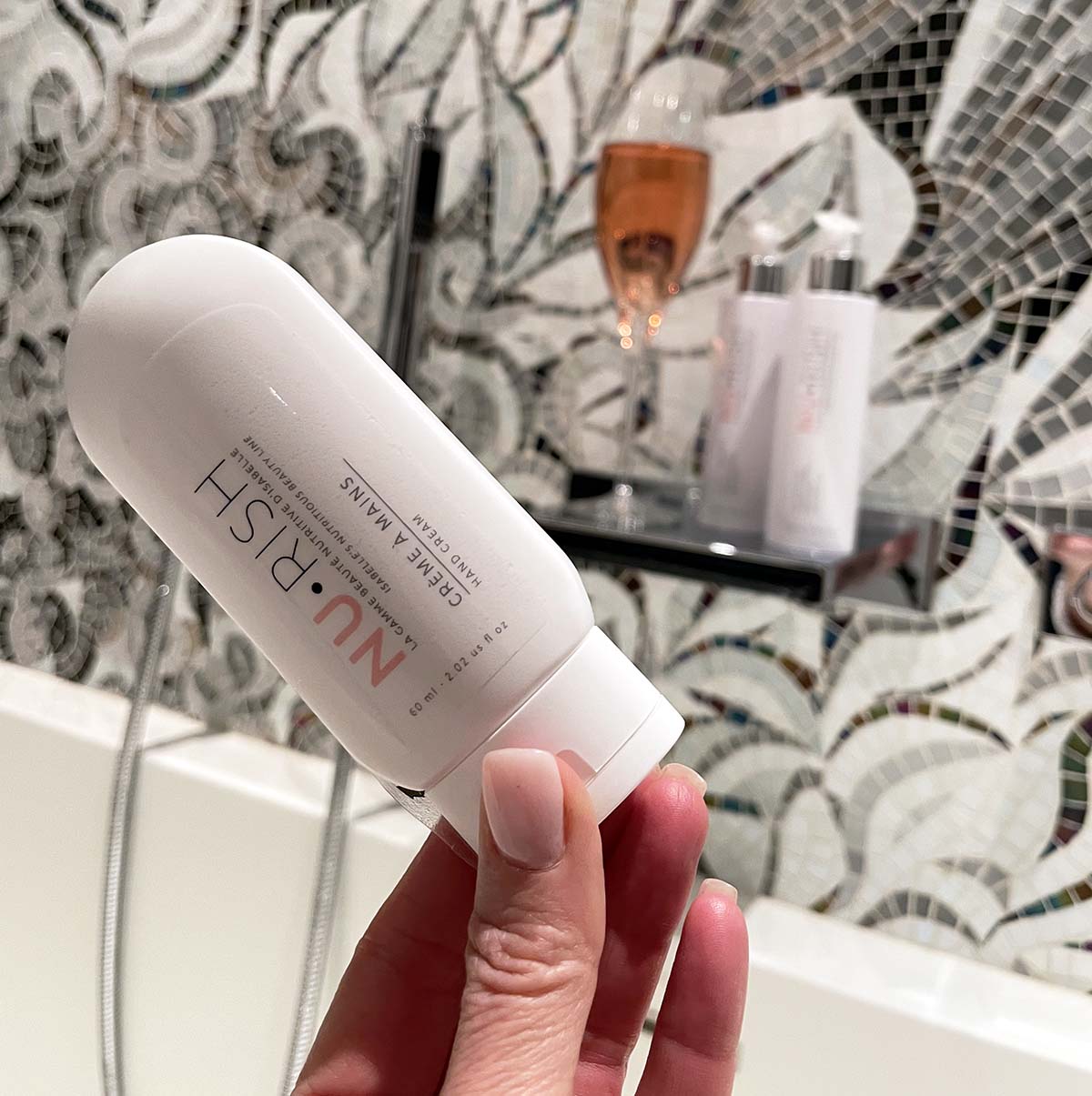 Photo de la crème à mains, composé à 98% d'ingrédients d'origine naturelle, végane, et non testés sur les animaux, tenu dans la main d'Isabelle Huot, devant une baignoire. Photo of the hand cream, composed of 98% of ingredients of natural origin, vegan, and not tested on animals, in the hand of Isabelle Huot next to a bathtub.