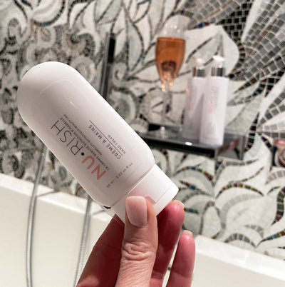 Photo de la crème à mains, composé à 98% d'ingrédients d'origine naturelle, végane, et non testés sur les animaux, tenu dans la main d'Isabelle Huot, devant une baignoire. Photo of the hand cream, composed of 98% of ingredients of natural origin, vegan, and not tested on animals, in the hand of Isabelle Huot next to a bathtub.