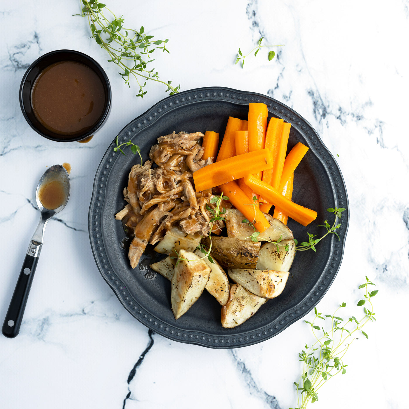 Déposé sur un comptoir de marbre, cette assiette est remplie de bâtonnets de carottes, de quartiers de pommes de terre assaisonnés et d'une généreuse portion de porc effiloché. La sauce du porc effiloché est déposé aux côtés de l'assiette principal. / Placed on a marble counter, this plate is filled with carrot sticks, seasoned potato wedges and a generous portion of pulled pork. The pulled pork sauce is placed alongside the main course.